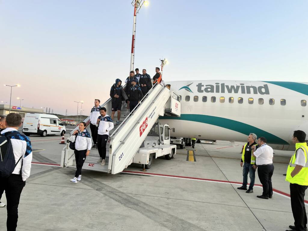 L'équipe de football du Luxembourg descendant de l'avion de la compagnie Tailwind. L'avion est blanc avec des bandes bleu. Sur le tarmac de l'aéroport d'Istanbul, on aperçoit le staff de l'aéroport et le pilote.