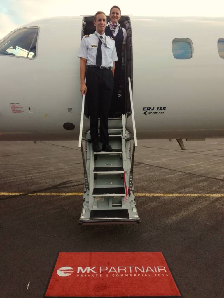 Captain and flight attendant smiling as they welcome passengers at the top of the airstair