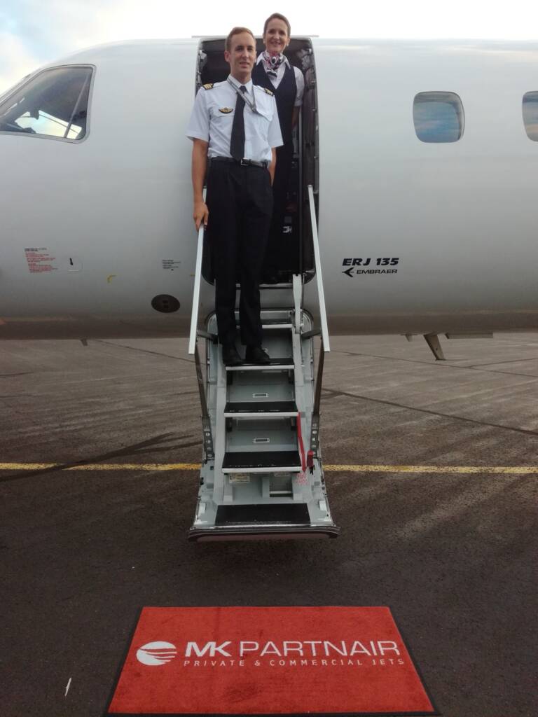 Commandant et hôtesse souriants sur la passerelle, tapis MK Partnair au premier plan