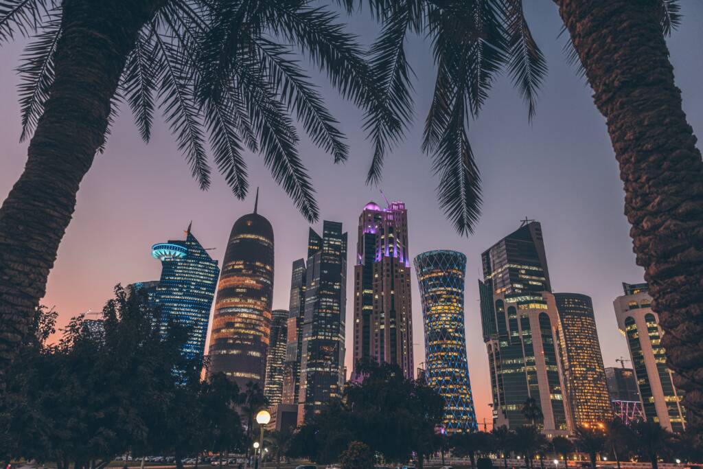Image montrant les buildings dans la ville de Doha, c'est le soir et il y a des palmiers et beaucoup de lumières sur les buildings